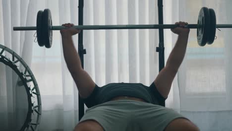 Man-doing-bench-press-workout-with-a-barbell-in-the-gym-by-a-window-with-a-trampoline