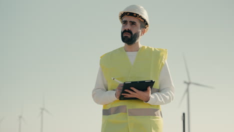 Auditing-wind-turbines-in-a-field-of-renewable-energy-generators-on-a-sunny-day,-a-professional-engineer-in-a-white-helmet-and-reflective-vest-uses-a-tablet-with-technology-to-promote-sustainability