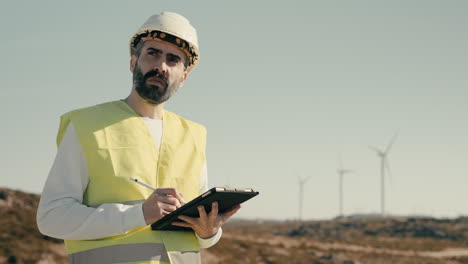 Promoting-clean-energy-and-sustainability,-a-professional-engineer-in-a-white-helmet-and-reflective-vest-uses-a-tablet-to-audit-wind-turbines-on-a-sunny-day