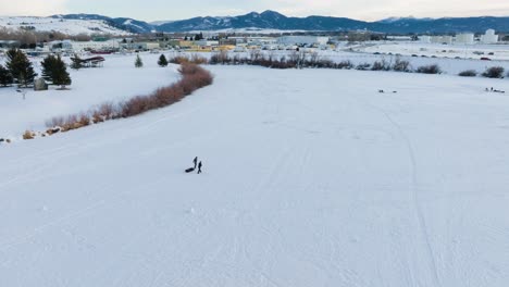 Boseman-Montana-Winter-Aeria-over-sledders-and-ice-fishermen-in-snowy-suburban-park,-orbit-right-with-4k-drone-over-with-mountain-backdrop-at-sunset