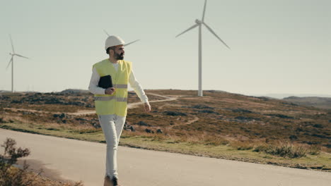 Ensuring-a-sustainable-future-for-our-planet,-a-professional-engineer-in-a-white-helmet-and-reflective-vest-uses-a-tablet-to-audit-wind-turbines-in-a-field-of-clean-energy-generators-on-a-sunny-day