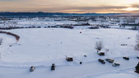 Boseman-Montana-Aerial-Winter-sunset-over-snowy-suburban-park,-orbit-right-with-4k-drone-over-ice-fishermen-with-mountain-backdrop