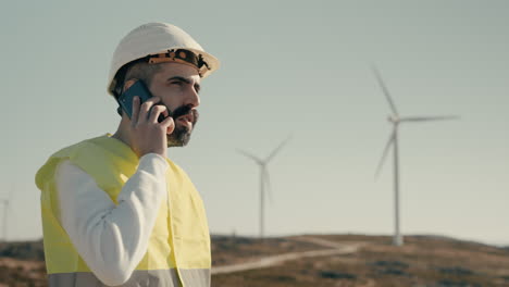 Close-up-of-a-Caucasian-engineer-in-a-field-of-wind-turbines,-answering-a-phone-call-on-a-sunny-day