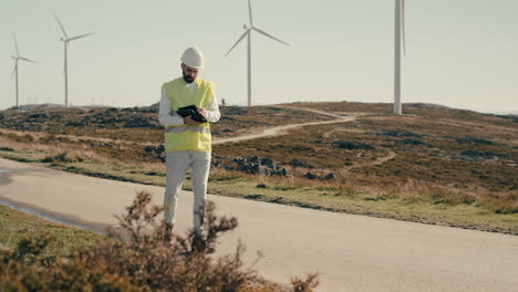 A-wide-shot-of-a-renewable-energy-engineer-in-a-reflective-vest-walking-on-a-field-of-renewable-energy-generators,-using-a-tablet-to-inspect-wind-turbines,-showcasing-the-role-of-technology
