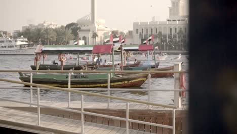 Holzboote-Mit-Emiratischer-Flagge-Liegen-Im-Leerlauf-Im-Dubai-Creek,-Während-Andere-Boote-Im-Hintergrund-Vorbeifahren