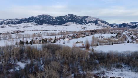 Boseman-Montana-Aerial-Winter-tilt-down-over-snowy-suburban-neighborhood-homes,-4k-drone-over-ice-fishermen-and-snow-park-with-mountain-backdrop