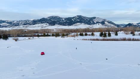 Boseman-Montana-Aerial-flythrough-over-snowy-suburban-park-at-golden-hour,-4k-drone-over-ice-fishermen-and-sledders-with-mountain-backdrop