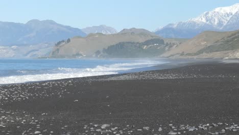 Sea-spray-hangs-in-air-before-low-hills-and-a-mountain-range-on-a-mid-winter's-day---Black-Sand-Beach,-Kaikoura-Coastline