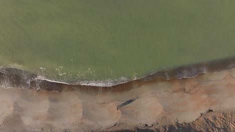Aerial-view-Two-people-slowly-walk-across-the-sand,-and-sea-waves-are-washing-the-shore-of-the-beach-on-a-cold-autumn-morning