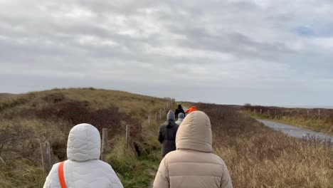 Rear-view-showing-group-of-people-walking-between-dunes-during-cloudy-day-in-slow-motion-during-cold-and-windy-day