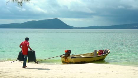 Man-carries-two-small-suitcases-to-small-boat-on-the-shore-of-a-tropical-beach