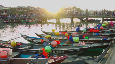 Cinematic-view-of-foot-bridge-with-people-walking-over-river-with-traditional-boats-with-riders-floating-on-the-river-at-sunset-in-old-town-of-Hoi-an,-Vietnam