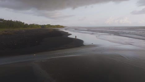 Frau-Läuft-Am-Atemberaubenden-Schwarzen-Sandstrand-Von-Bali,-Filmische-Luftaufnahme-–-Fernweh