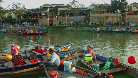 Sampan-Bootsfahrer-Warten-In-Ihren-Booten-Auf-Kunden-Im-Fluss-Thu-Bon-Der-Antiken-Stadt-Hoi-An-Mit-Traditionellen-Bambushäusern-Im-Hintergrund,-Vietnam