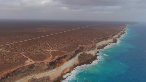 Aerial-view-showing-Van-on-empty-road-above-gigantic-Cliffs-and-ocean-during-cloudy-day---Nullarbor-SA,-Australia