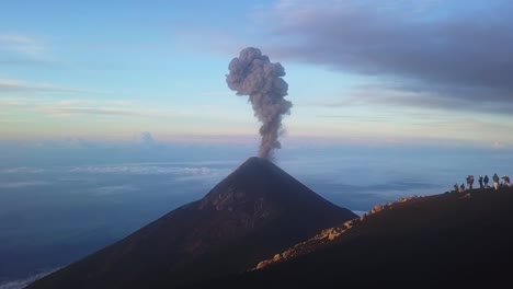 Volcán-En-Erupción-Al-Amanecer-Con-Gente-Parada-En-Una-Colina-Mirándolo