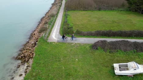 People-riding-bicycles-on-a-turn-near-a-boat-and-ocean-in-the-village,-Aerial-orbit-shot