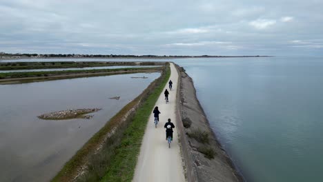 Family-riding-bicycles-at-Loix-village-in-Île-de-Ré-Island-in-Western-France-while-toddler-holds-mother's-hand,-Aerial-follow-behind-shot