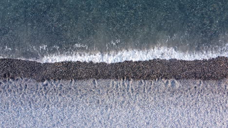 Top-down-aerial-view-above-layered-white-sand-and-pebble-beach-washed-over-by-foaming-ocean-waves