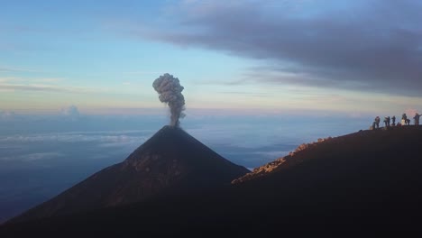 Ein-Vulkan-Bricht-Im-Sonnenaufgang-Aus,-Während-Eine-Gruppe-Von-Menschen-Von-Einem-Anderen-Vulkan-Aus-Zusieht