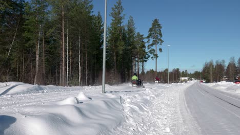 Two-snowmobiles-driving-on-the-side-of-the-road,-with-forest-on-the-left-side,-on-cold-sunny-winter-day-in-Sweden