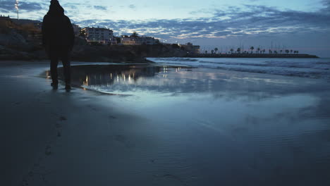 Man-footprints-wet-sand-at-winter-beach-blue-hour,-twilight-with-coastal-city