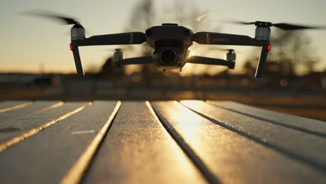 Black-Mavic-2-on-table-outdoors-starting-up-and-taking-off,-with-a-blurred-sunset-and-country-background