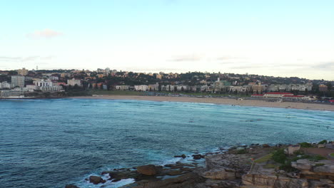 Drone-flying-across-Bondi-bay-towards-the-beach-as-people-paddle-on-the-water-in,-Sydney-Australia