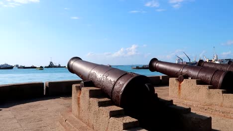 Panning-shot-of-old-rusted-cannons-with-boats-navigating-in-the-port-of-Forodhani