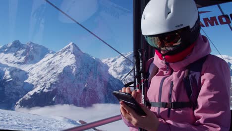 Woman-in-ski-suit-texting-on-smartphone-inside-a-cable-car-of-a-mountain-ski-resort-in-sunny-day