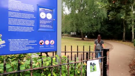 Panning-shot-of-the-Holland-Park-information-sign-with-a-young-woman-walking-through-the-park