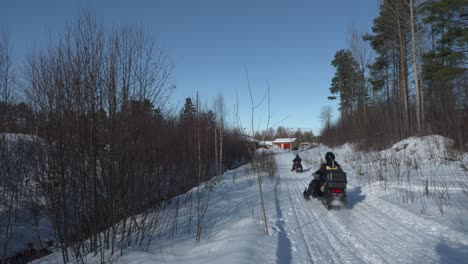 Two-snowmobiles-driving-on-a-path-with-trees-on-both-side,-on-cold-sunny-winter-day-in-Sweden