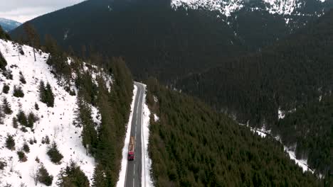 Aerial-drone-flight-of-truck-loaded-with-wood-driving-through-the-snow-covered-mountains-and-forests-on-a-road-in-Transylvania-Romania-Europe-2023---Timber-transport---bird-view
