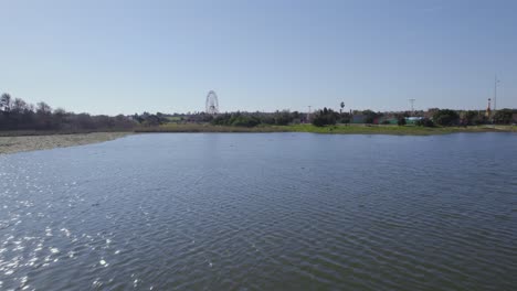 Flying-over-an-artificial-lake-with-grass-around,-and-a-small-theme-park-with-a-Ferris-wheel-in-the-background