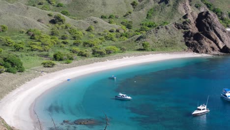 White-sandy-beach-at-secluded-bay-with-rugged-mountains-and-boats-moored-in-calm,-turquoise-blue-green-ocean-water-of-Padar-Island,-East-Nusa-Tenggara-of-Indonesia