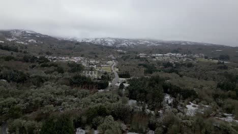 Snowy-village-with-vegetation-in-winter-and-snowy-mountains-on-the-horizon-under-a-cloudy-sky-in-Picornio,-in-Galicia