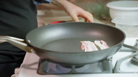 Person-with-an-arm-tattoo-placing-half-slices-of-bacon-in-a-Teflon-frying-pan-on-a-gas-stove