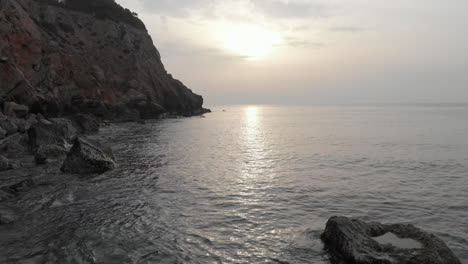as-sun-rises-on-horizon-of-calm-sea,-low-tracking-drone-shot-glides-along-rocky-coastline