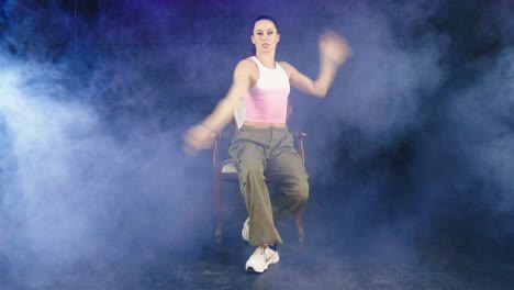 Young-Caucasian-female-contemporary-professional-dancer-performing-sitting-on-chair-in-smoky-studio-room