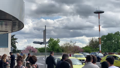 People-waiting-at-taxi-airport-pick-up-lane-while-huge-military-airplane-approaches-ground-in-cloudy-background
