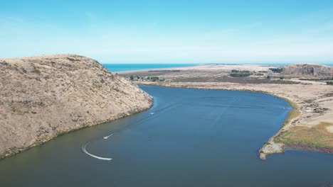 Aerial-view-of-deep-blue-lake,-coastline,-volcanic-peninsula-and-power-boats-racing-around-course---Lake-Forsyth
