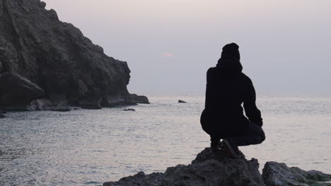 man-sitting-on-rough-rock-watching-sunrise-as-seagulls-fly-over-sea,-fascinated-by-beautiful-view