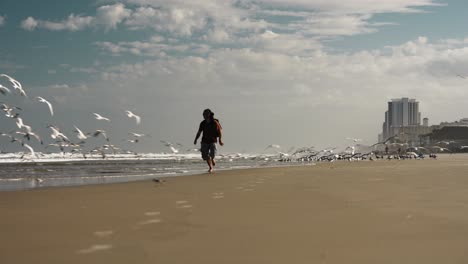 Epische-Szene-Eines-Mannes,-Der-Am-Strand-Läuft,-Während-Tausende-Von-Möwen-Fliegen