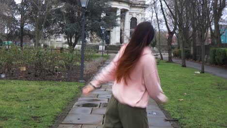 Long-haired-stylish-female-urban-dancing-on-park-pavement-towards-white-building-with-columns