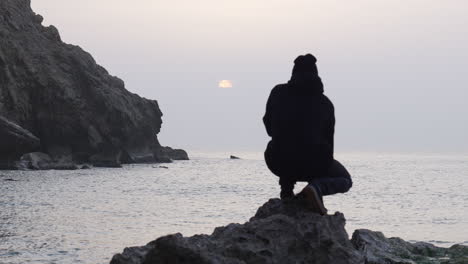 man-jumps-onto-a-rough-rock-and-sits-down-to-watch-sunrise-as-seagulls-fly-over-sea,-fascinated-by-beautiful-view