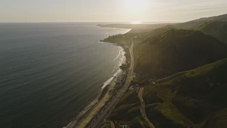 Aerial-drone-shot-flies-right-from-a-Malibu-sunset-over-Pacific-Coast-Highway-PCH-to-a-green-mountain-at-golden-hour
