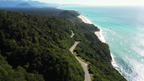 New-Zealand-aerial-view-of-sunny-weather-in-west-coastline-with-asphalted-road-following-the-coastline-of-the-blue-ocean-water-in-green-lush-forest