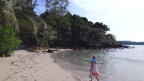 Elderly-lady-walking-barefoot-on-white-beach-in-summer-dress
