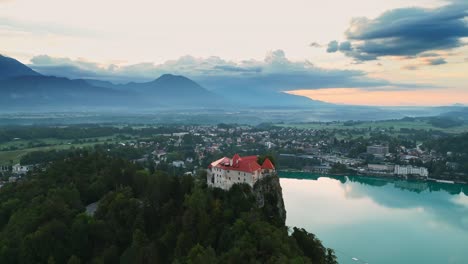 Bled-Castle-On-Rock-Overlooking-Beautiful-Lake-Bled-Slovenia-And-Town-With-Mountains-in-Background,-Aerial-Parallax-Drone-Shot,-Peaceful-Travel-Destination,-Early-Morning-Sunrise,-Cinematic-4K