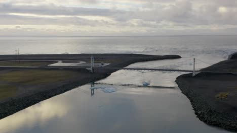Aerial-backwards-shot-of-Icelandic-ocean,-bridge,-river-mouth-and-glacier-with-icebergs-floating-on-water-during-cloudy-day
